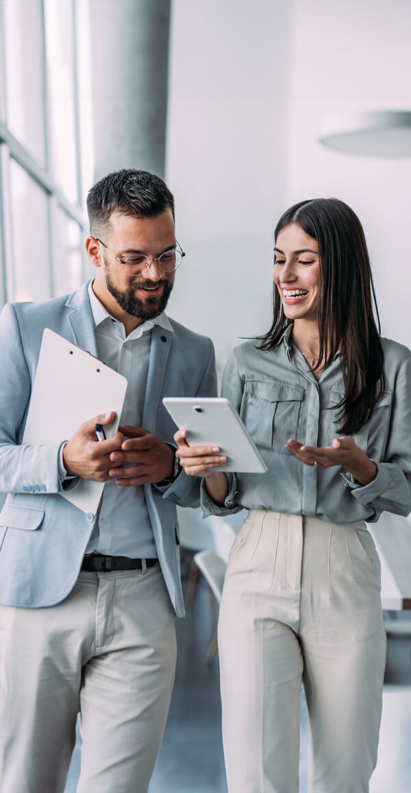 A man and woman in light business attire walk through a modern office, smiling and discussing something on a digital tablet.