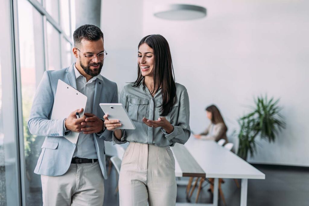 A man and woman in light business attire walk through a modern office, smiling and discussing something on a digital tablet.