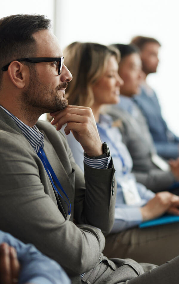 A row of attentive professionals sits in a conference setting, with a man in focus resting his chin on his hand and listening intently.