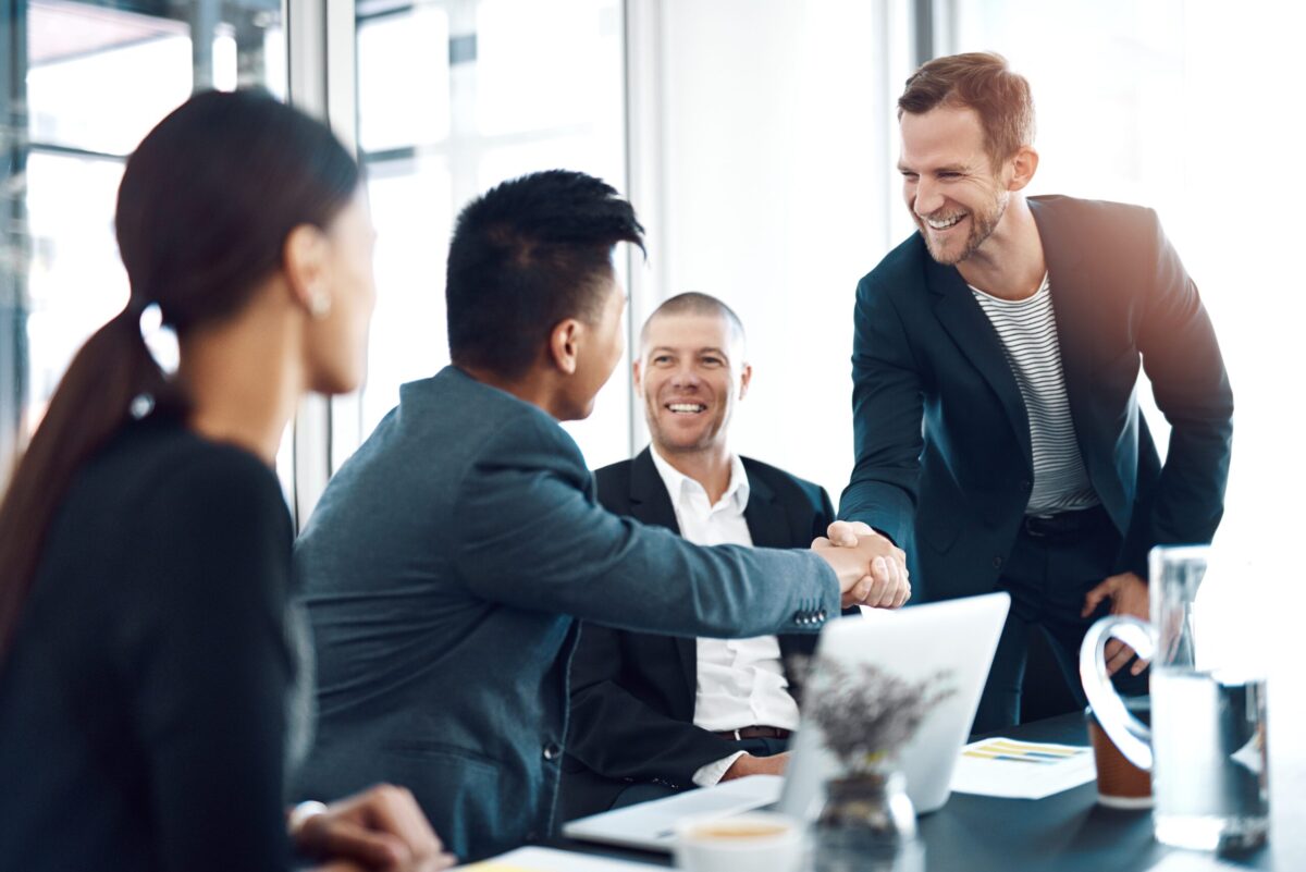 Men and women sitting at an office desk, wearing business suit jackets and shirts. 2 men shaking hands.