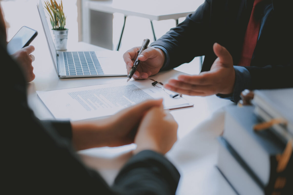 View of two people's hands, people sitting at desk, one hand holds a pen. Papers on table. Business meeting setting.